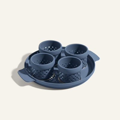The Egg Poacher features four blue perforated silicone cups on a matching handled tray, ideal for making poached eggs. Displayed against a plain background, it offers a convenient alternative to the Always Pan.