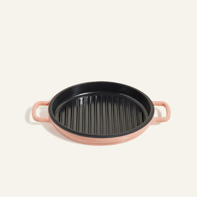 The Cast Iron Hot Grill is a round, pink enameled cast iron pan with ridges and two handles, displayed on a light background.
