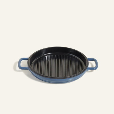 The Cast Iron Hot Grill is a round blue enameled grill pan with dual handles and raised ridges on the cooking surface, displayed against a light background.
