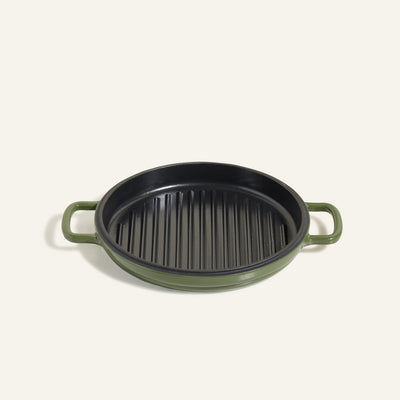 The Cast Iron Hot Grill is a round, green enameled cast iron grill pan with two handles and a ridged black surface—ideal for indoor grilling, shown against a plain light background.