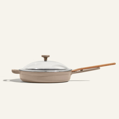 The Cast Iron Always Pan, a beige enameled nonstick frying pan with a wooden handle and glass lid, is photographed against a plain light background.