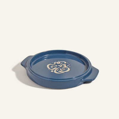 The Flipping Platter is a blue ceramic baking dish with handles and a cream floral design in the center, perfect for serving dishes like tahdig. Shown against a plain light background.