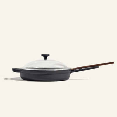 The Cast Iron Always Pan, featuring a black enamel finish, glass lid, and long wooden handle, is shown on a plain light background and offers 8-in-1 versatility for all your cooking needs.
