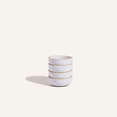 Four white Tiny Bowls, noted for their scratch-resistant ceramic finish, are stacked on a plain light background with a soft left shadow. These durable bowls are ideal as stylish and space-saving stackable tableware.