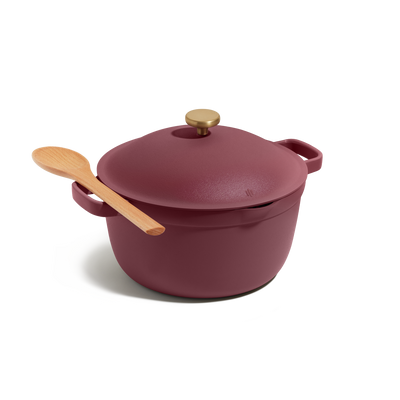 The Perfect Pot, a maroon cast iron Dutch oven with a gold knob, is displayed on a white background with a wooden spoon resting on its lid. This versatile pot is ideal for family meals.