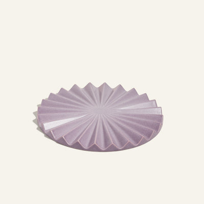 The Pleat Trivet is a round lavender ceramic dish with a geometric pleated design, ideal as a decorative kitchen accessory or sunburst trivet, displayed against a plain light background.