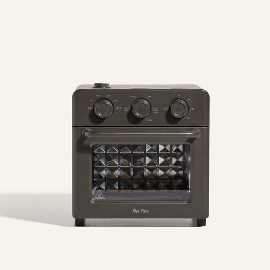 The AU - Wonder Oven is a dark gray countertop toaster oven with three top control dials and a front glass door featuring a diamond-patterned interior. "Our Place" appears at the bottom of the door. Doubles as an air fryer for extra versatility.