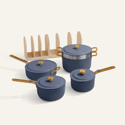 The Essentials Saucepan Set includes four blue pots and pans with wooden handles and lids, arranged beside a beige pan organizer rack on a light background. Features a nontoxic nonstick coating for easy cooking and cleaning.