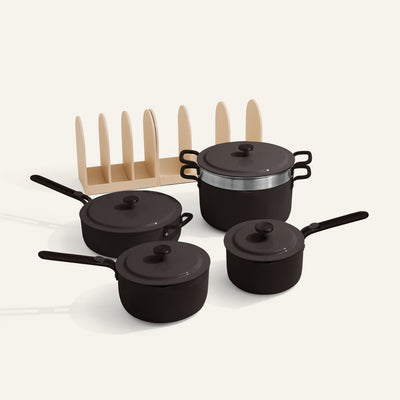 Four black pots and pans with lids from the Essentials Saucepan Set are placed next to a beige vertical organizer on a light background.