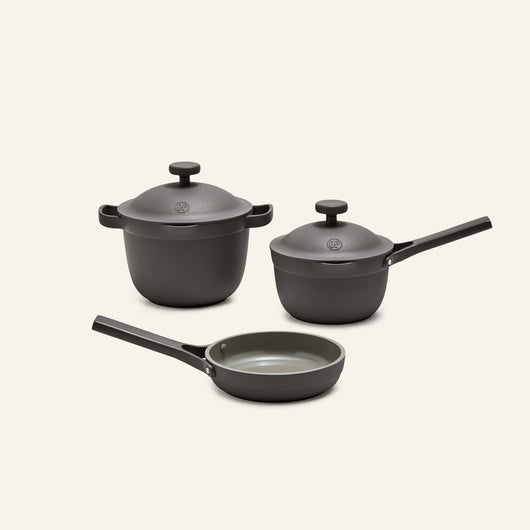 The Tiny Cookware Duo + Petite Perfect Pot set features a matte gray lidded pot, a lidded saucepan, and a frying pan with black handles, showcased on a simple light background.