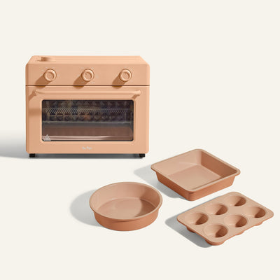 The Large Wonder Oven + Wonder Oven Baker's Kit in peach includes a countertop oven with three control knobs, shown with matching square, round, and muffin pans on a light background.
