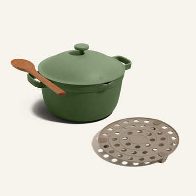 The Perfect Pot Roasting Rack and Perfect Pot (green) with a lid, plus a wooden spoon on the handle, sit beside a round beige trivet with holes.