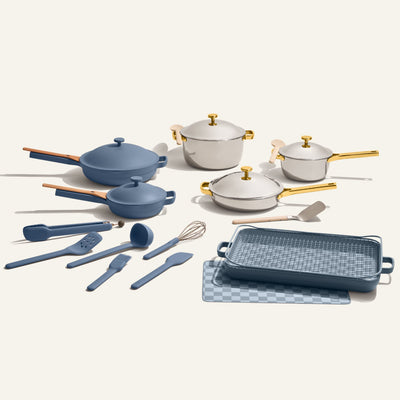 The Home Chef Set (25 piece) features non-toxic blue and white cookware and utensils—including pots, pans, spatulas, a whisk, and a griddle pan—with gold and wooden handles, all displayed on a neutral background.