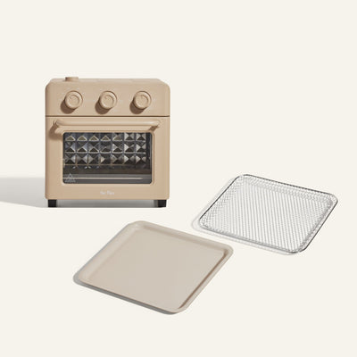 The Wonder Oven + Wonder Oven Essentials Kit is displayed in beige, featuring three dials, a glass door, and comes with a metal wire tray and solid baking tray beside it on a white background.