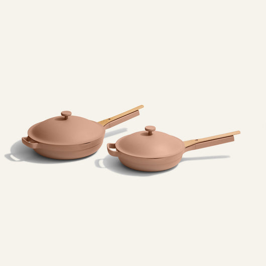The Always Pan Duo, featuring two tan pans with lids and wooden handles in different sizes, sits side by side on a white surface—highlighting versatile cookware for any kitchen.