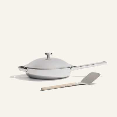 The Titanium always pan pro non toxic frying pan, a white lidded frying pan with a long handle and innovative nonstick technology, sits next to a metal spatula with a beige handle on a light background.