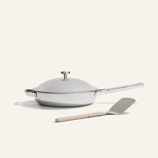 The Titanium always pan pro non toxic frying pan, a white lidded frying pan with a long handle and innovative nonstick technology, sits next to a metal spatula with a beige handle on a light background.