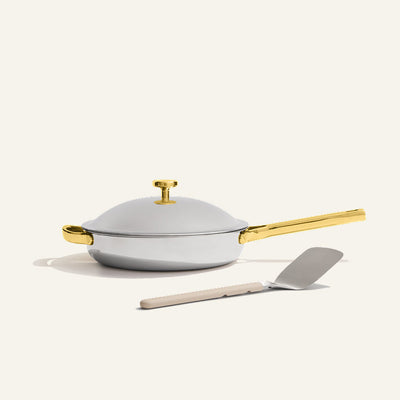 The Titanium always pan pro non toxic frying pan, a white no-coating nonstick frying pan with a gold handle, sits on a light surface with a metal spatula placed in front.
