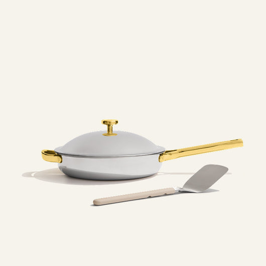 The Titanium always pan pro non toxic frying pan, a white no-coating nonstick frying pan with a gold handle, sits on a light surface with a metal spatula placed in front.