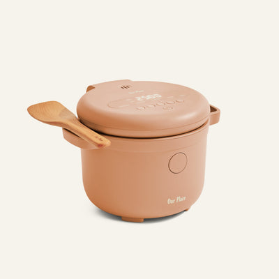 The Rice Cooker by Our Place, in beige, features fuzzy logic, a digital display, and comes with a wooden spoon resting on its handle for perfect rice preparation, all set against a simple backdrop.