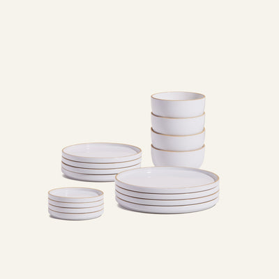 The Complete Plates & Bowls Set features handcrafted ceramic dinnerware with tan rims, including stacked bowls, small plates, and large plates, beautifully arranged on a light background.