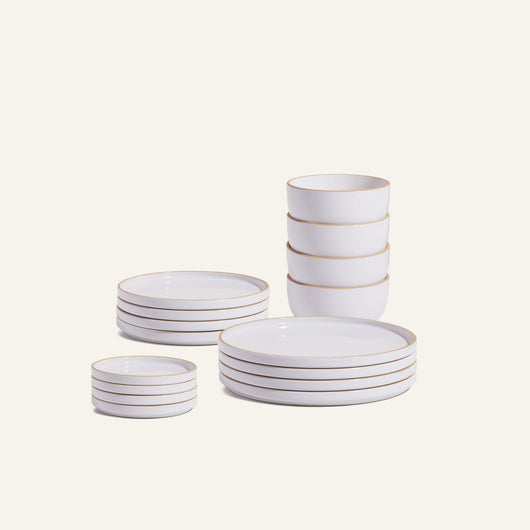 The Complete Plates & Bowls Set features handcrafted ceramic dinnerware with tan rims, including stacked bowls, small plates, and large plates, beautifully arranged on a light background.