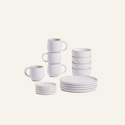 The Dinnerware Set features white ceramic plates, bowls, mugs, and small dishes with light brown rims, all displayed on a plain light background.