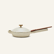 A cream-colored ceramic frying pan with a matching lid and a reddish-brown wooden handle, shown on a solid off-white background.