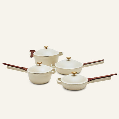 The Cookware Set features four cream-colored pieces with wooden handles and gold knobs: two pans, an non toxic ceramic nonstick always pan saucepan with lid, and a pot with lid, all displayed on a light background.