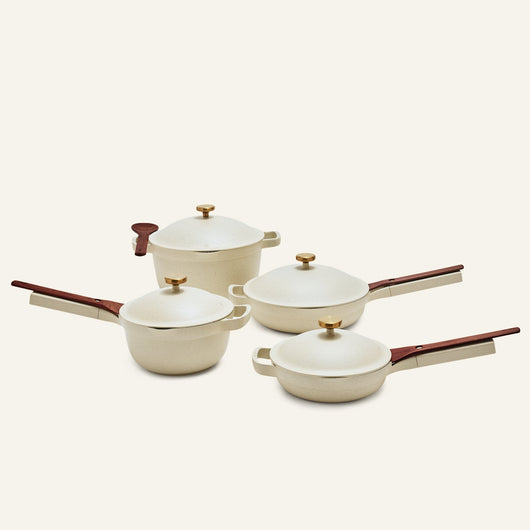 The Cookware Set features four cream-colored pieces with wooden handles and gold knobs: two pans, an non toxic ceramic nonstick always pan saucepan with lid, and a pot with lid, all displayed on a light background.