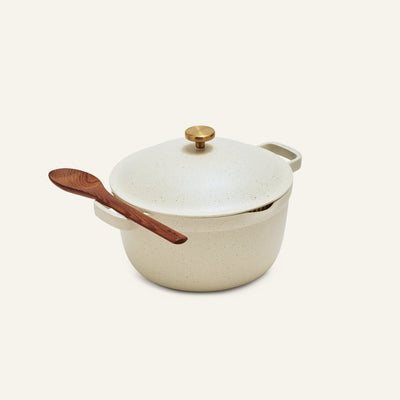 The perfect pot non toxic nonstick dutch oven, a cream ceramic family-sized pot with a lid and gold knob, features a wooden spoon resting on its handle, shown against a plain light background.